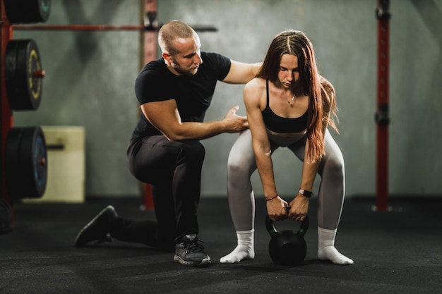Mulher jovem musculosa malhando com personal trainer na academia. Treinador auxiliando a mulher no treinamento com kettlebell.