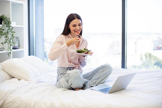 Mulher jovem na cama em casa e usando laptop moderno enquanto come salada saudável