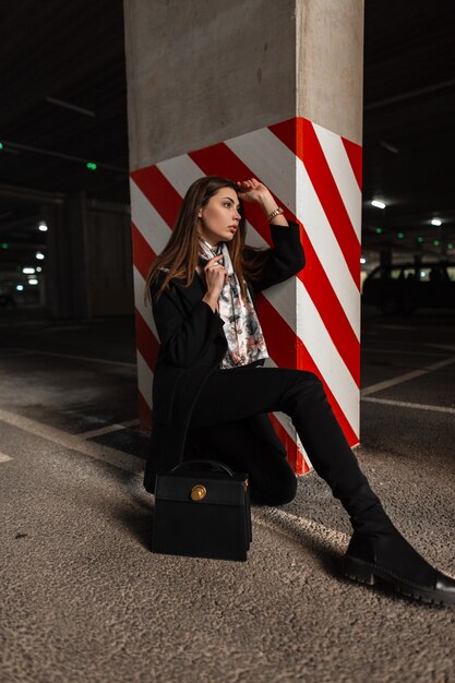 Mulher jovem na moda em elegante casaco preto com lenço de seda elegante em botas com bolsa de couro senta-se no asfalto no estacionamento perto do pilar em linhas vermelho-brancas. Modelo de moda garota atraente descansa.