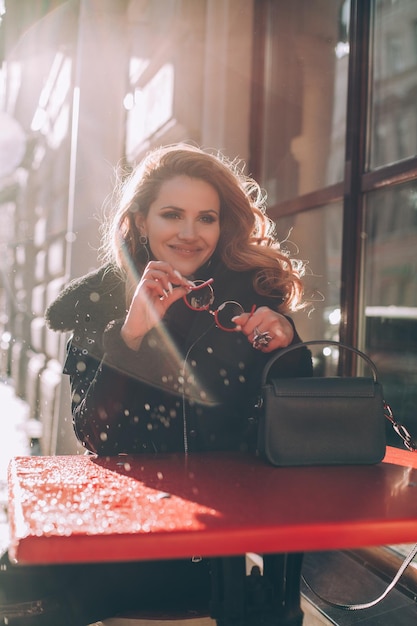 Mulher jovem na moda em óculos de sol vermelhos de jaqueta preta e bolsa azul Mulher café de rua Estilo de rua