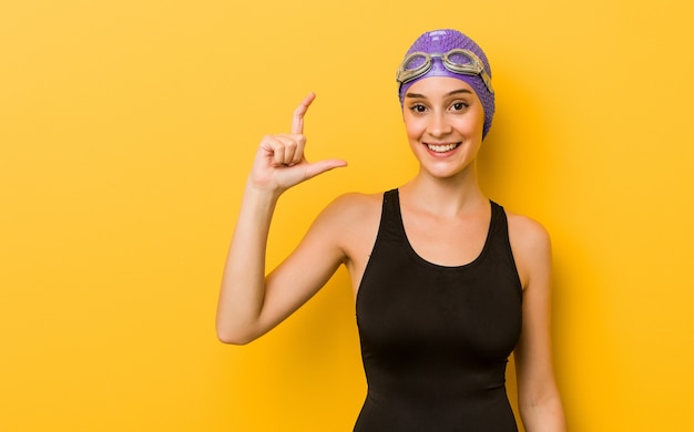 Mulher jovem nadadora caucasiana segurando algo pequeno com os indicadores, sorrindo e confiante.