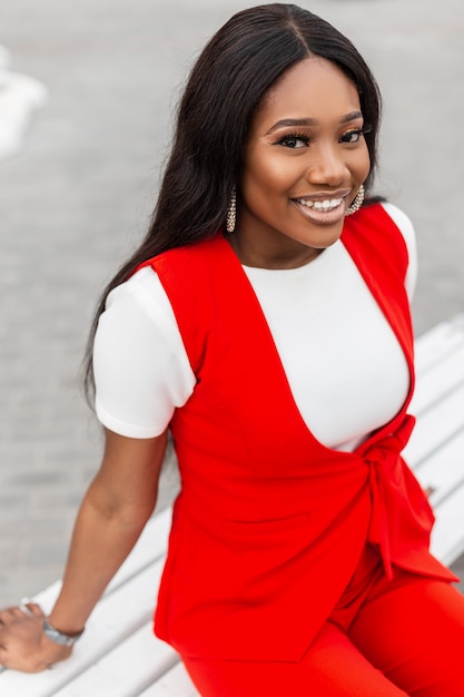 Mulher jovem negra muito positiva com um lindo sorriso com pele limpa e saudável em elegante t-shirt branca em colete vintage vermelho senta-se e sorri perto da cidade. rua retrato alegre jovem africana.