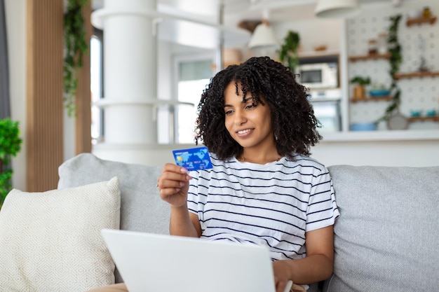 Mulher jovem no sofá comprando on-line com cartão de débito Bela mulher africana usando computador portátil para compras on-line em casa