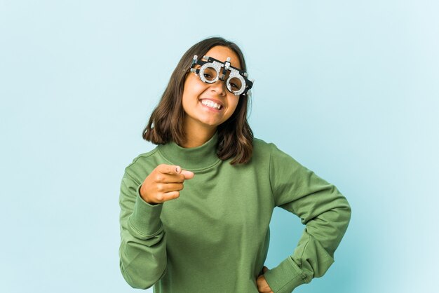 Mulher jovem oculista latina sobre sorrisos alegres de parede isolada apontando para frente.