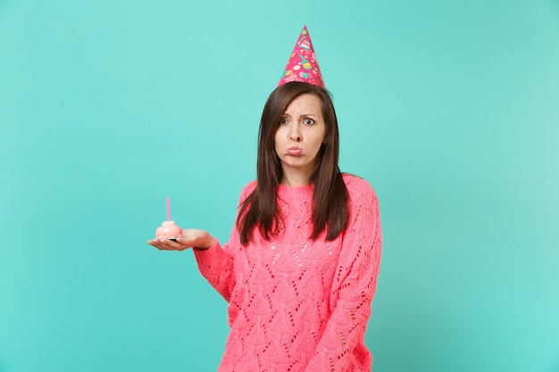 Mulher jovem ofendida na camisola de malha rosa e chapéu de aniversário, segurando na mão o bolo com vela isolada no retrato de estúdio de fundo de parede azul turquesa. Conceito de estilo de vida de pessoas. Simule o espaço da cópia.