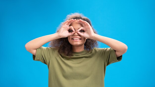 Mulher jovem olhando positivamente para a frente usando uma camiseta verde-oliva com binóculos isolados na parede azul