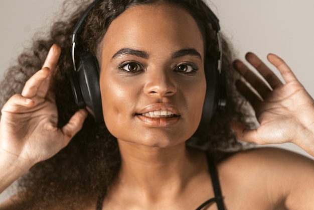 Mulher jovem ouvindo música em fones de ouvido, com uma blusa preta isolada em uma parede cinza.