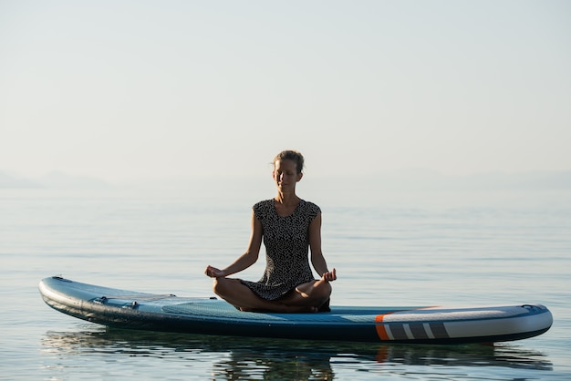 Mulher jovem pacífica com uma expressão serena no rosto, meditando em posição de lótus em uma prancha de supino flutuando na água.