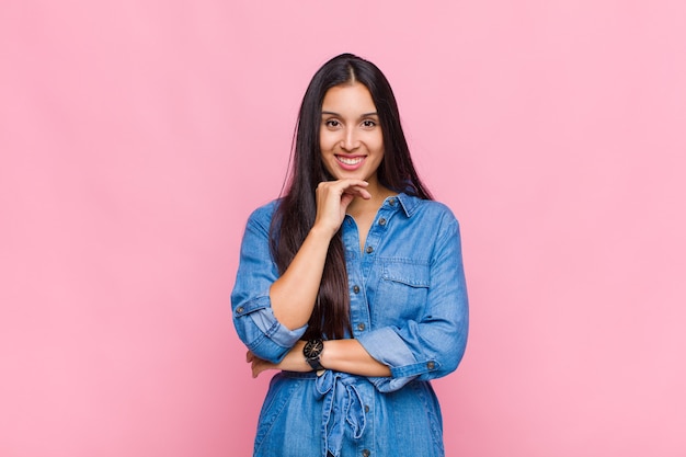 Mulher jovem parecendo feliz e sorrindo com a mão no queixo, pensando ou fazendo uma pergunta