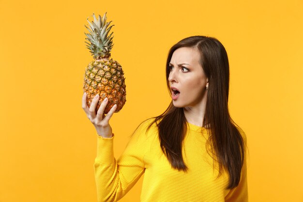 Mulher jovem perplexa chocada em roupas casuais segurando, olhando em frutas frescas de abacaxi maduras isoladas em fundo amarelo laranja. Estilo de vida vívido de pessoas, relaxe o conceito de férias. Espaço de cópia de mock up