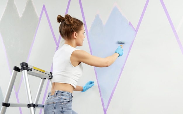 Mulher jovem pintando a parede com rolo de pintura e usando fita adesiva em pé na escada