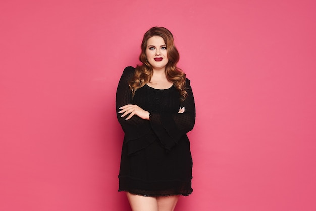 Mulher jovem plus size modelo caucasiana em vestido preto com as mãos postas e posando sobre uma parede rosa isolada com espaço de cópia