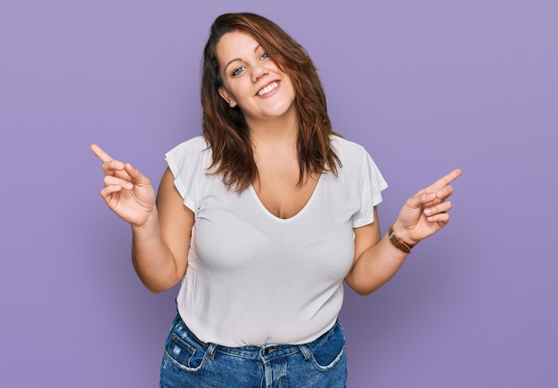 Mulher jovem plus size vestindo camiseta branca casual sorrindo confiante apontando com os dedos para diferentes direções copie o espaço para propaganda