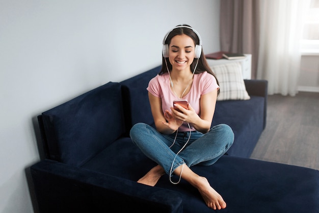 Mulher jovem positiva e feliz sorrindo. Ela se senta no sofá na sala. Modelo ouve música. Ela posa com as pernas cruzadas. Jovem mulher segurar o telefone nas mãos.