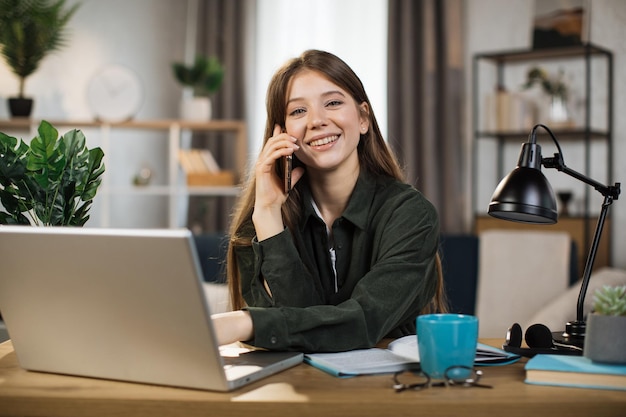 Mulher jovem positiva falando no celular e trabalhando no laptop no escritório brilhante