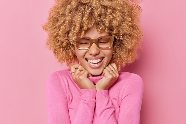 Mulher jovem positiva feliz sorri amplamente se sente divertido mantém as mãos na gola do jumper lembra de algo engraçado usa óculos fica divertido isolado sobre fundo rosa. conceito de emoções.