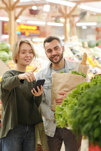Mulher jovem positiva usando aplicativo de smartphone para comprar e apontar verduras para o namorado enquanto eles compram produtos no mercado