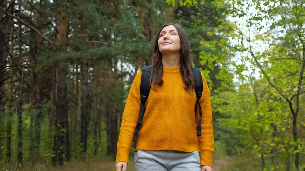 Mulher jovem, positiva, vestindo um suéter laranja elegante, caminha ao longo de uma estrada rural em meio a uma floresta verde com pinheiros antigos no fim de semana no dia de outono