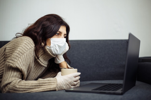 Foto mulher jovem que trabalha em casa durante a quarentena devido à pandemia de coronavírus. linda garota fica em casa usando luvas e máscara médica e digitando em um laptop. conceito mundial de epidemia covid-19.