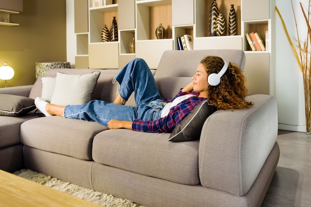 Mulher jovem relaxando ouvindo música em casa reclinada em um sofá da sala de estar com um sorriso de prazer