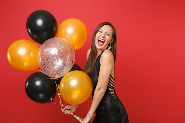 Mulher jovem rindo em um vestido preto comemorando segurando balões de ar isolados sobre fundo vermelho. Dia internacional da mulher de São Valentim feliz ano novo, conceito de festa de feriado de maquete de aniversário.