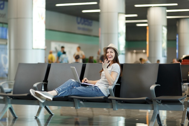 Mulher jovem rindo viajante turista sentada, trabalhando em um laptop, espalhando as mãos e esperando no saguão do aeroporto internacional