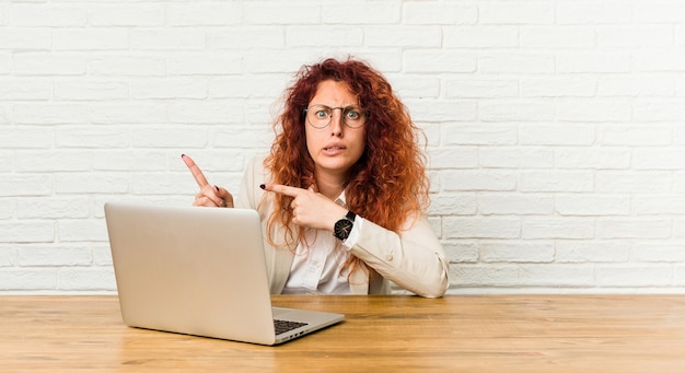 Mulher jovem ruiva cacheada trabalhando com seu laptop chocada apontando com o dedo indicador para um espaço de cópia.