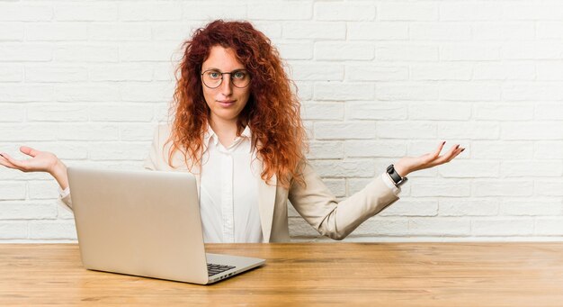 Mulher jovem ruiva cacheada trabalhando com seu laptop, mostrando uma expressão de boas-vindas.