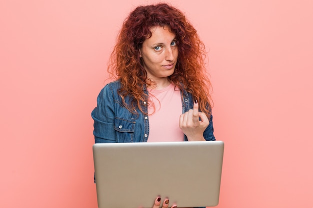 Foto mulher jovem ruiva caucasiano segurando um laptop apontando com o dedo para você, como se convidando se aproximar.