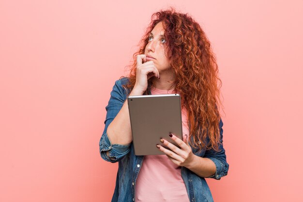 Mulher jovem ruiva caucasiano segurando um tablet olhando de soslaio com expressão duvidosa e cética.