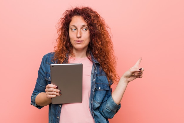 Mulher jovem ruiva caucasiano segurando um tablet sorrindo alegremente apontando com o dedo indicador.