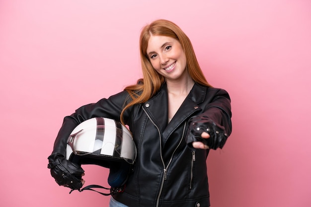 Mulher jovem ruiva com um capacete de moto isolado no fundo rosa, apontando para a frente com expressão feliz