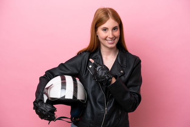 Mulher jovem ruiva com um capacete de moto isolado no fundo rosa, apontando para o lado para apresentar um produto