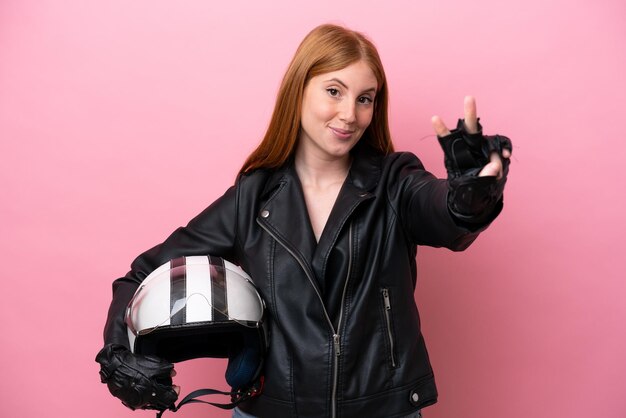 Mulher jovem ruiva com um capacete de moto isolado no fundo rosa sorrindo e mostrando sinal de vitória