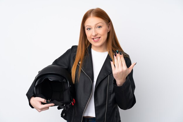 Mulher jovem ruiva com um capacete de moto sobre parede branca isolada, convidando para vir com a mão.