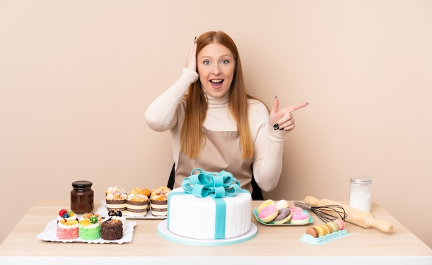 Mulher jovem ruiva com um grande bolo surpreso e apontando o dedo para o lado