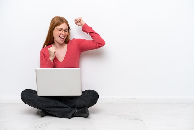 Mulher jovem ruiva com um laptop sentado no chão isolado no fundo branco comemorando uma vitória