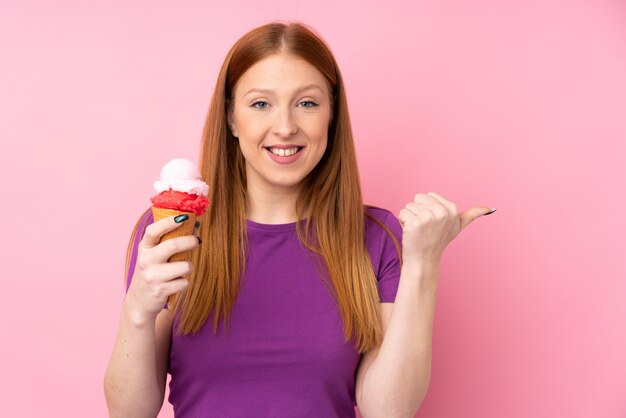 Mulher jovem ruiva com um sorvete de corneta sobre parede rosa isolada, apontando para o lado para apresentar um produto