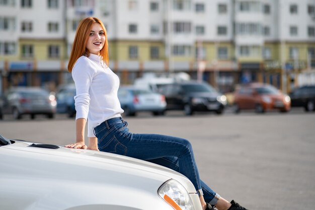 Mulher jovem ruiva elegante camisola branca, calça jeans azul elegante e tênis pretos, sentado em um capô do carro novo e caro na rua da cidade, aproveitando o dia quente de verão.