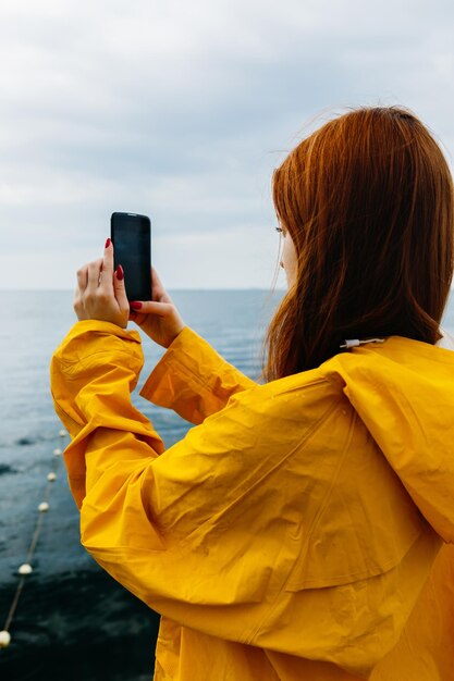 Mulher jovem ruiva em capa de chuva amarela posando na costa do oceano tirando foto da natureza com smartphone