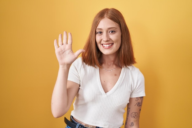 Mulher jovem ruiva em pé sobre fundo amarelo mostrando e apontando para cima com os dedos número cinco enquanto sorria confiante e feliz.