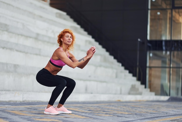 Mulher jovem ruiva europeia em roupas esportivas fazendo agachamentos ao ar livre