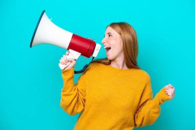 Mulher jovem ruiva isolada em fundo azul gritando através de um megafone para anunciar algo na posição lateral