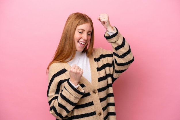 Mulher jovem ruiva isolada em fundo rosa comemorando uma vitória