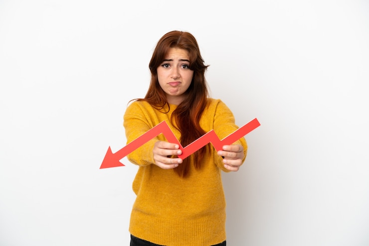 Foto mulher jovem ruiva isolada no fundo branco segurando uma seta para baixo e com uma expressão triste