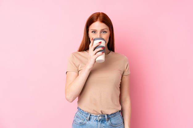 Mulher jovem ruiva isolada rosa segurando e tomando um café para levar