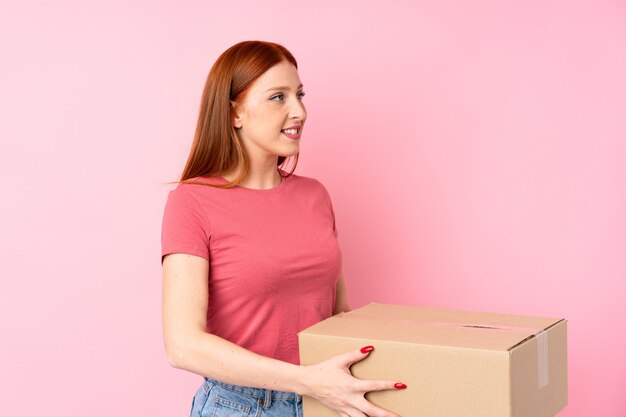 Mulher jovem ruiva isolado rosa segurando uma caixa para movê-lo para outro site