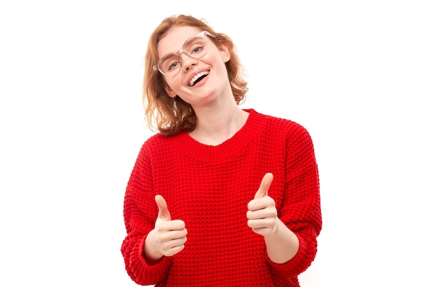 Mulher jovem ruiva mostra a mão com polegares para cima e sorri para a câmera isolada no fundo branco