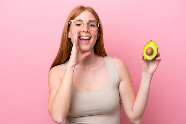 Mulher jovem ruiva segurando um abacate isolado no fundo rosa gritando com a boca aberta