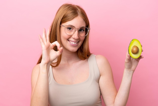Mulher jovem ruiva segurando um abacate isolado no fundo rosa, mostrando sinal de ok com os dedos